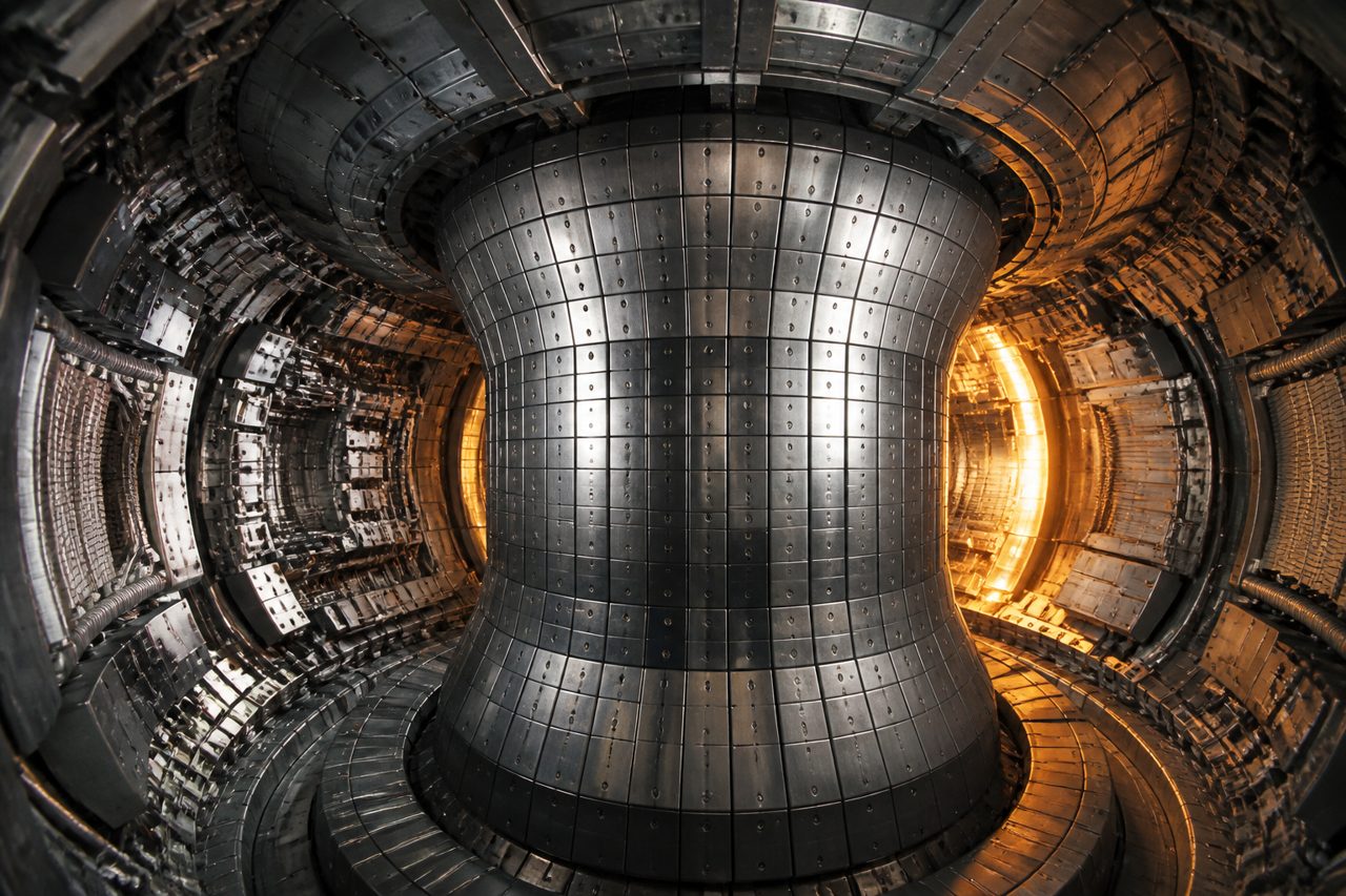 Inside a tokamak vessel: steel tiles curving around a central column, lit amber at the far wall.
